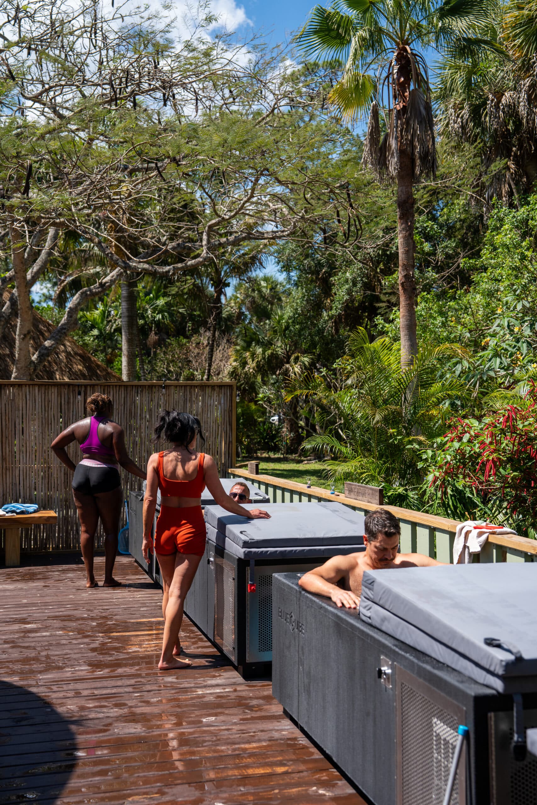 People in colorful swimsuits stand and lounge by large outdoor hot tubs on a wooden deck, with palm trees and lush greenery nearby.