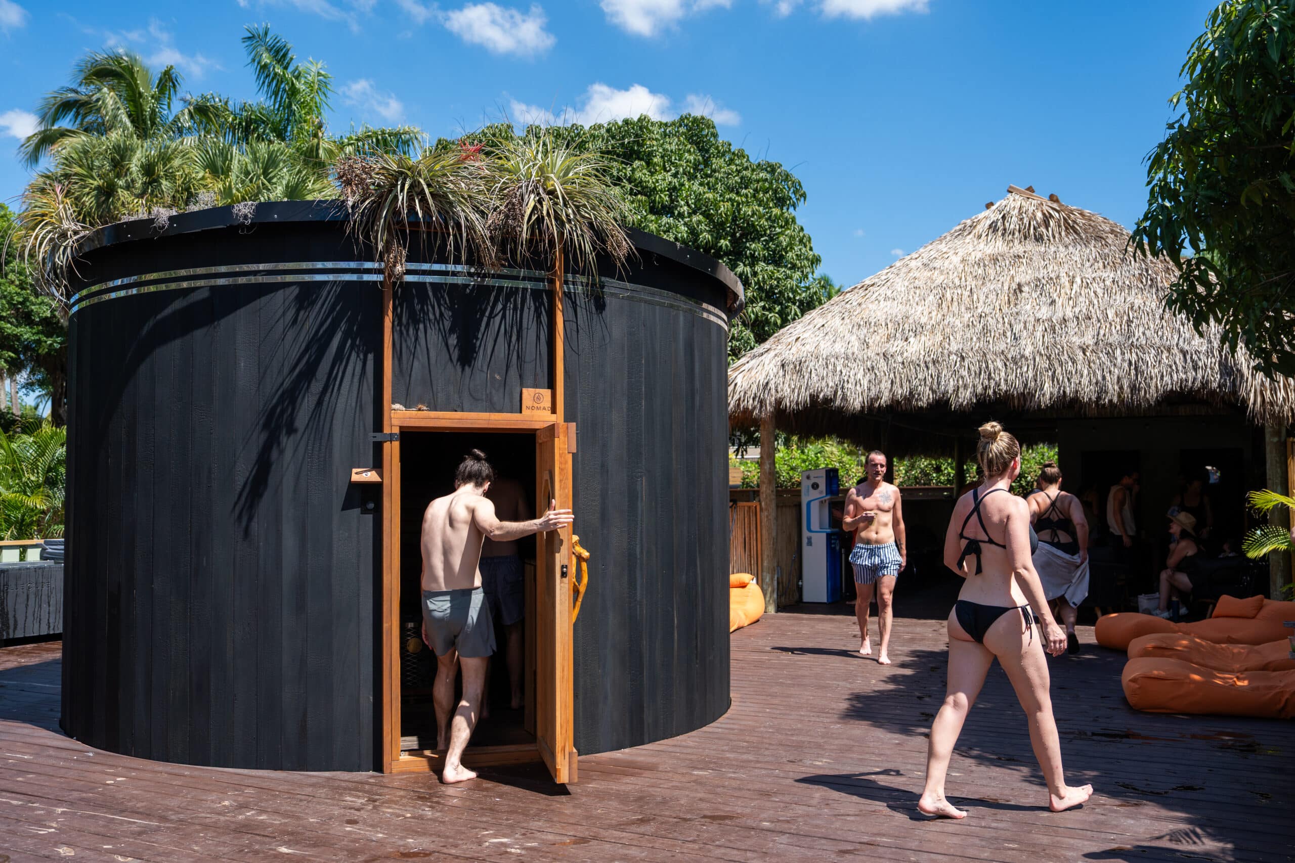People in swimsuits around a round black wooden hut with plants on top at a tropical outdoor resort.