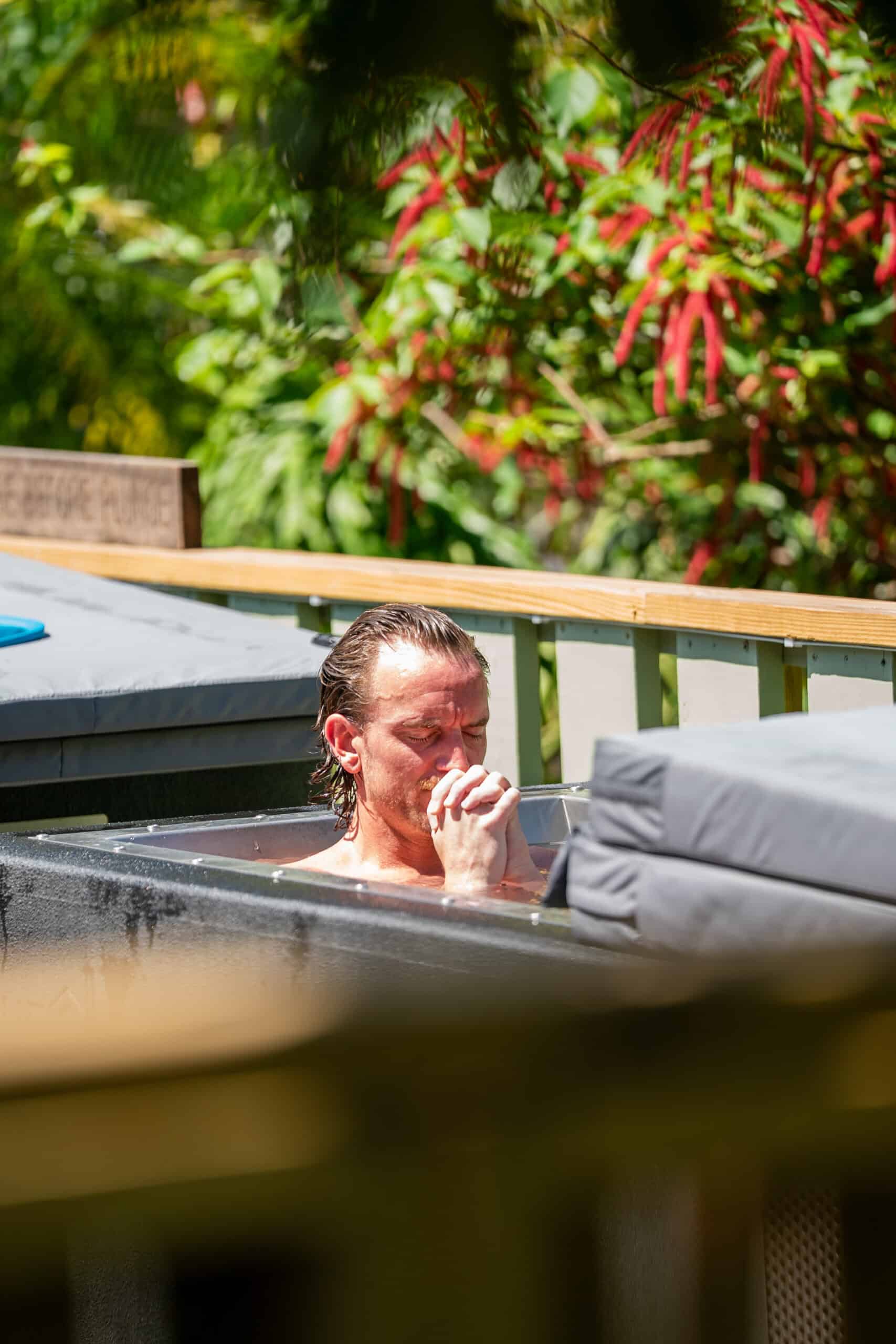 Man relaxing in an outdoor hot tub with hands near his face, wood railing and green plants in the background.