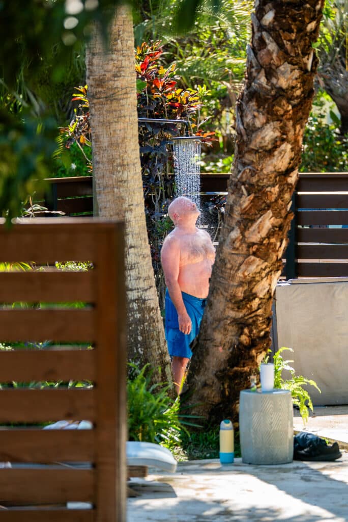 Man standing outdoors in blue shorts under a wall-mounted outdoor shower with water spraying, surrounded by palm trees and greenery.