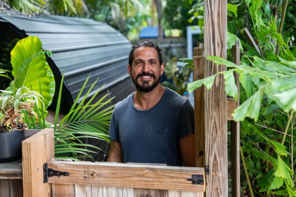 Smiling man with dark hair and beard standing behind a wooden crate in a lush garden setting.