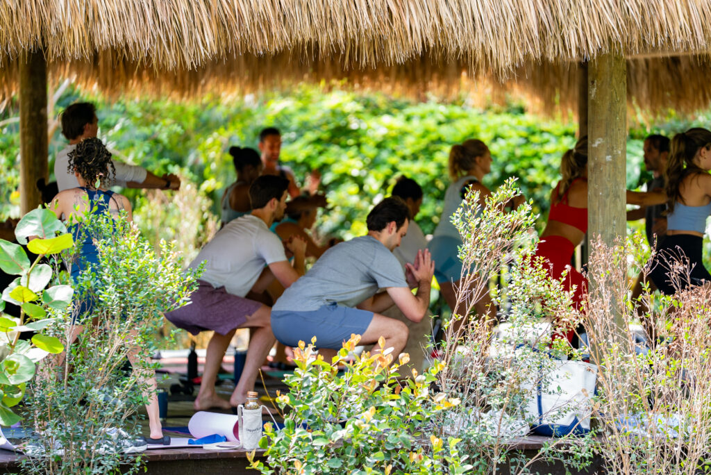 Group of people practicing yoga under a thatched shelter in a lush outdoor area.