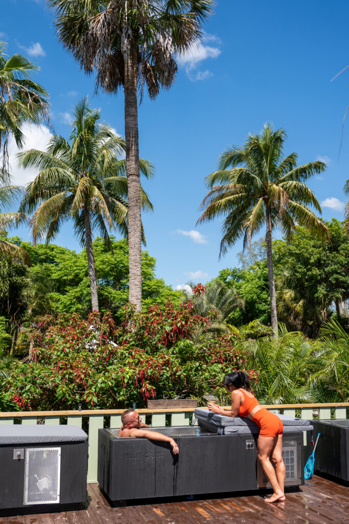 Outdoor pool area with dark hot tubs labeled Blue Cube, a man reclining in one tub and a woman in an orange swimsuit leaning over another tub, surrounded by tall palm trees against a bright blue sky.