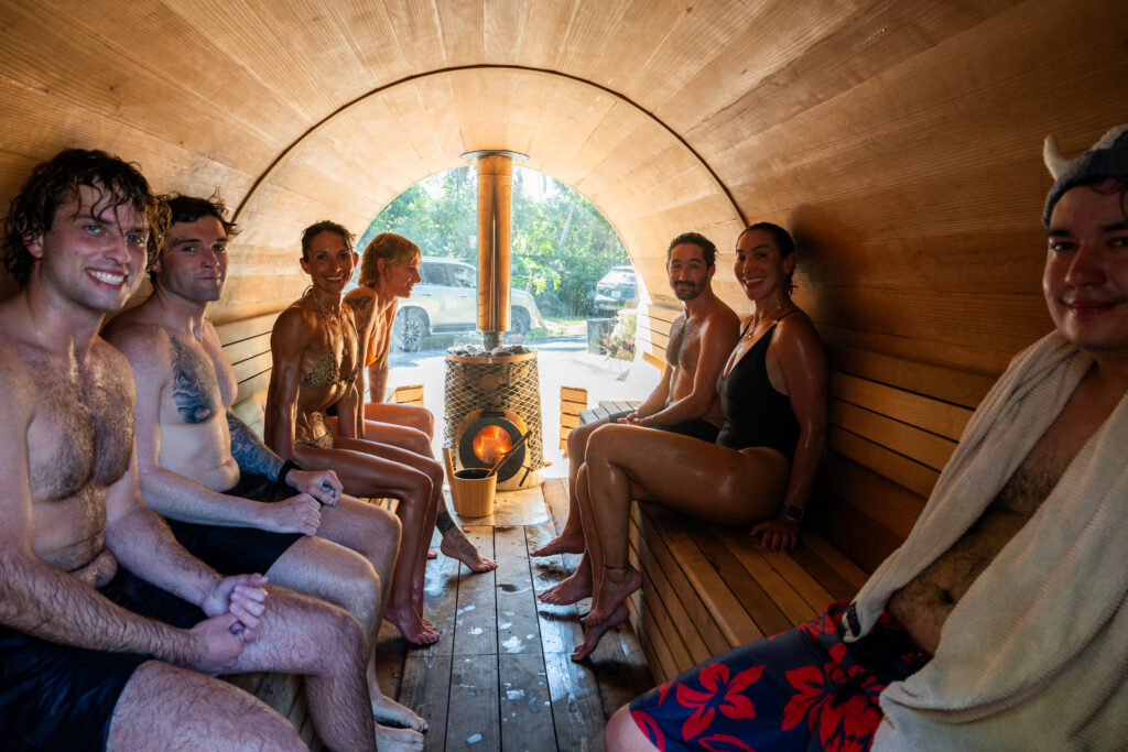 Eight adults in a cedar sauna, wearing swimsuits, seated along wooden benches with a central heater highlighted by outdoor light.