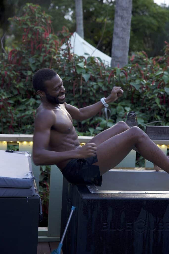 Man in dark swim trunks sits shirtless in a pool or hot tub, laughing with a clenched fist raised outdoors.
