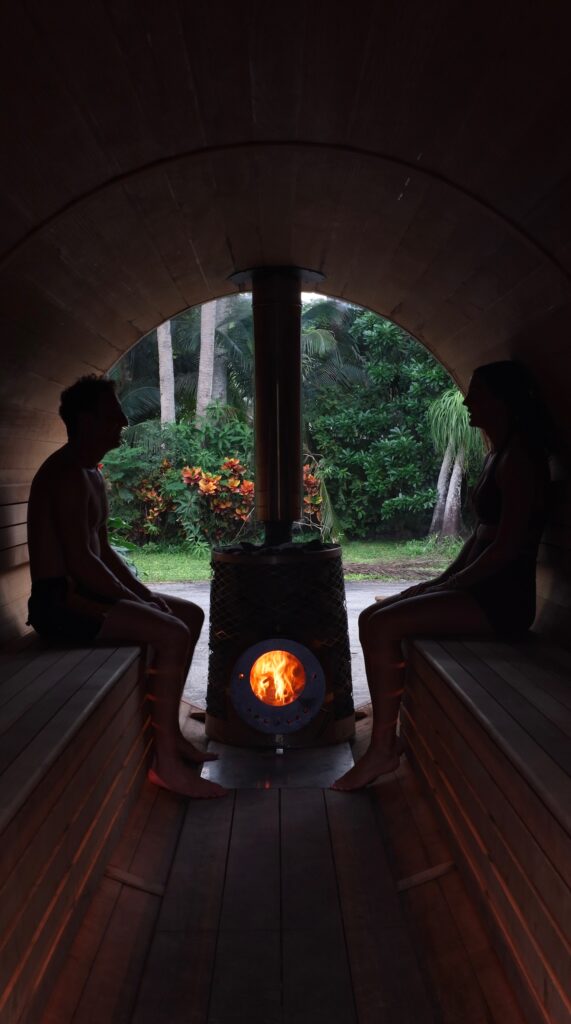 Two people sit on opposite benches inside a round wooden sauna, facing a central wood stove with a fire, opening to a lush garden outside.